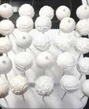 A display of white cake pops on sticks, decorated with smooth icing, swirls, dots, and small pearl-like accents, arranged on a white stand.