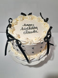 Vintage style cake A round birthday cake with white frosting, decorated with black ribbon bows, small black hearts, and the text 'happy birthday Claudz' written on top, shown on a white plate against a plain light background.