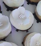 A top-down view of several frosted cupcakes arranged on a white surface. Each cupcake is topped with piped white frosting and a large white flower decoration with a small pearl-like center.