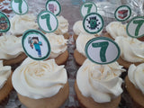A close-up of several frosted cupcakes arranged in a tray, each topped with a round white pick with a green border, a large green number 7, and a gaming-themed character or block graphic.