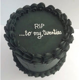 A round black cake with piped ruffled frosting borders and white icing text on top that reads 'RIP to my twenties .