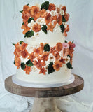 A two-tier white wedding or celebration cake decorated with orange, peach, and white flowers and green leaves, displayed on a round wooden cake stand against a white fabric backdrop.
