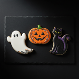 Three Halloween-themed decorated cookies shaped like a white ghost, an orange jack-o'-lantern, and a black cat with purple tail details, arranged on a dark slate serving board against a black background.