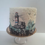 A custom lighthouse cake decorated with textured white frosting, a hand-painted lighthouse and house, and blue-green waves at the base, displayed on a clear glass cake stand.