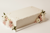 A rectangular cream-colored cake with smooth frosting, decorated with pink and white roses and green leaves along the lower left and right sides, displayed on a white cake board against a clean white background.