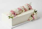 A rectangular white cake decorated with pink and white flowers and green leaves, displayed on a clean white background.
