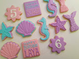 A set of decorated sugar cookies shaped like seashells, a seahorse, starfish, and a mermaid tail, with pastel pink, purple, and turquoise icing on a light cream background. Two cookies feature birthday messages: 'HAPPY BIRTHDAY SCARLETT' and 'HAPPY BIRTHDAY LAYA'.