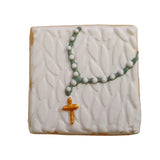A close-up of decorated sugar cookies with white icing, featuring a central square cookie with a leaf pattern and a small gold cross, surrounded by other iced cookies on a white surface.