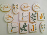 A set of decorated sugar cookies arranged on a white background. The cookies are round and rectangular, decorated with white icing and gold, green, and brown designs including a cross, a dove, a necklace, leaves, and the letter J.