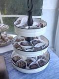 A three-tier white and black serving stand filled with chocolate-covered pastries and iced treats, set on a blue tablecloth in a bright indoor setting.