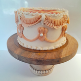 A decorated round cake with beige and peach frosting, piped ruffles, and pearl-like accents, displayed on a round wooden cake stand against a plain light background.