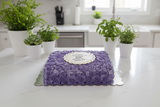 Purple floral cake on a white surface with potted plants in the background