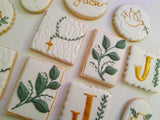 Decorated sugar cookies with white icing, featuring green leaf designs, gold monogram letters, and a green rosary motif on a light background.