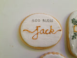 A round white-frosted cookie with 'GOD BLESS Jack' written in icing, shown on a plain light background with other decorated cookies partially visible at the edges.